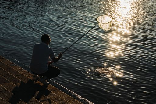 A man on the shore crouching holding a fishnet above the lake, and a reflection of sunset sunlight on the surface