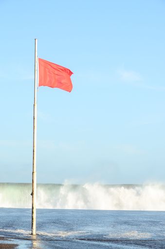 A vertical shot of a red flag on the shore with waves in the background.  Atlantic Ocean