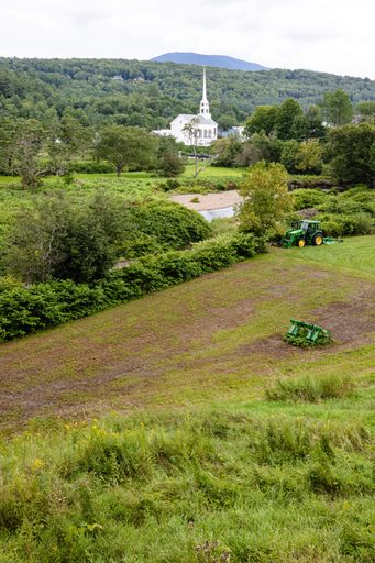 A vertical of a tractor cultivating farmland against Stowe Community Church on green hills of Vermont