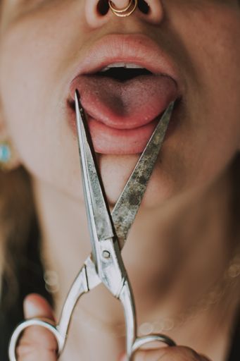 A vertical shot of a caucasian girl with nose rings holding scissors over her tongue