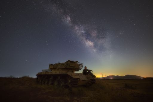 A man stargazing while sitting on a rusty tank with mountains and lights in the background