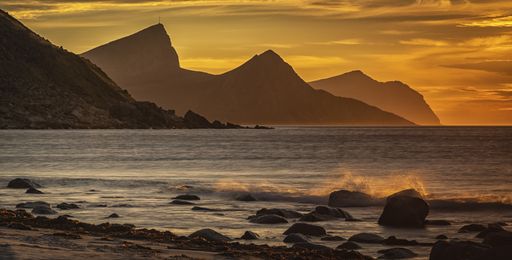 A beautiful shot of the sea waves hitting the rocks on the shore with mountains and a yellow sky in the background