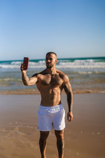 A vertical shot of an attractive topless man with tattoos standing  on a beach and taking selfies with his phone