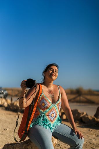A vertical shot of an attractive woman with tattoos sitting on a rock with a camera in her hand