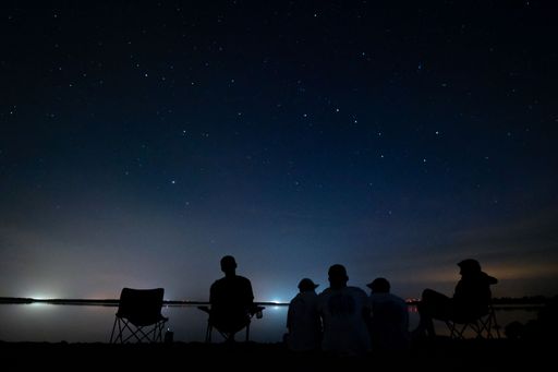 A group of people sitting on the coast of river with starry sky at night in the background