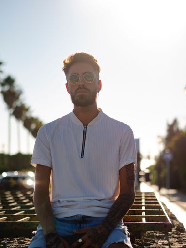 A vertical shot of a young sexy tattooed male with facial hair and sunglasses posing near palm trees