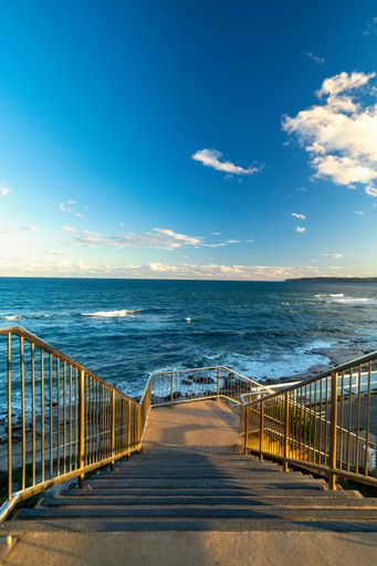 A descending wooden stairs with fences on blue sea under clue cloudy sky, vertical shot