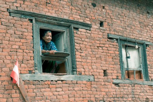 An old female looking out from the open window of an old brick house