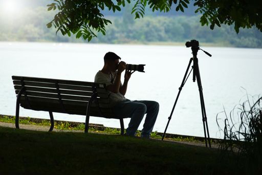A young male sitting on a bench on the shore of lake taking photo with a professional camera