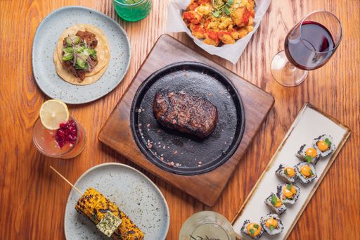 A flat lay of different variations of food with beef steak, sushi, wine, and others on a wooden table