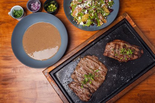 A flat lay of beef steak, gravy, and a bowl of salad on a wooden table