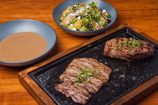 A flat lay of beef steak, gravy, and a bowl of salad on a wooden table