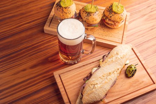 A flat lay of a beer mug full of beer with different types of sandwiches around on a wooden table
