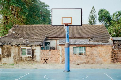 An old brick house behind a street basketball court and the hoop with big trees in the background