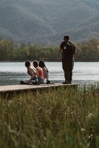 A vertical shot of three girls sitting on a pier and a man fishing in the lake against a forest