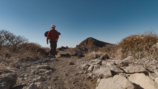 A man hiking along a narrow trail surrounded by a rocky wilderness in Japan under a clear sky