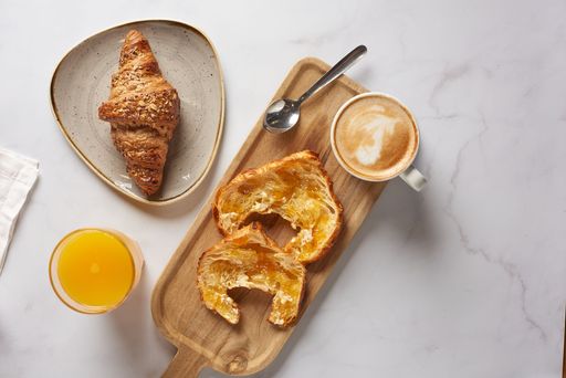 A French croissant with coffee and small spoon in wooden board with juice on marble surface
