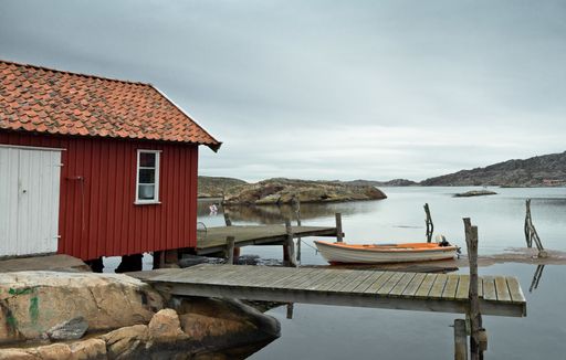 A red cabin on a wooden pier and a boat in the middle of a lake surrounded by rocky hills under the cloudy sky