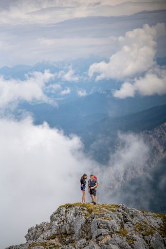A vertical shot of two young hikers standing on the rock in the mountains against white fluffy clouds
