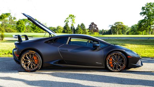 A black Lamborghini Huracan from the side with an open trunk parked in the road next to a park