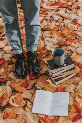 A vertical shot of a person wearing leather boots standing next to some books and a mug on the ground covered with autumn leaves
