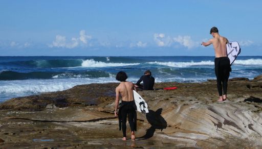 rear view of three young men carrying surfboards walking on sandstone rock towards watergoing for a surf