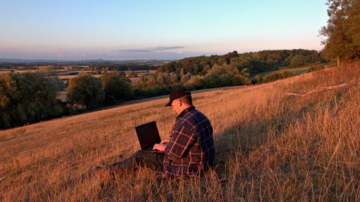 The view of a man using his laptop while sitting on a hill slope in the countryside