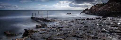 A panoramic shot of the sea with long exposure surrounded by rocks under a cloudy sky