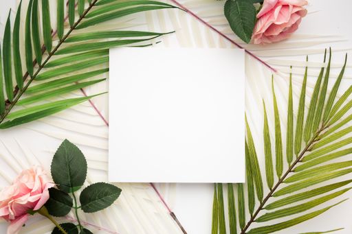 A top view of a blank square paper surrounded by palm leaves and roses isolated on a white background