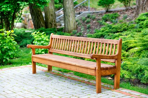 A beautiful and elegant handmade oak bench stands in a park on paving slabs with green bushes and large trees to the rear