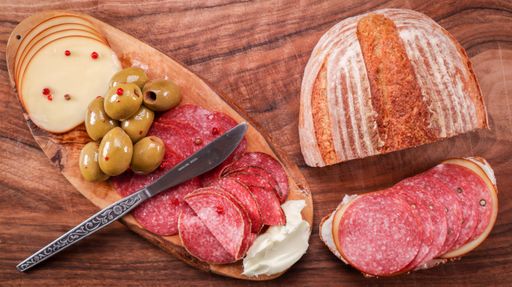 A top view of freshly baked bread on a wooden table next to a cutting board with sliced bread, sausage, and olives