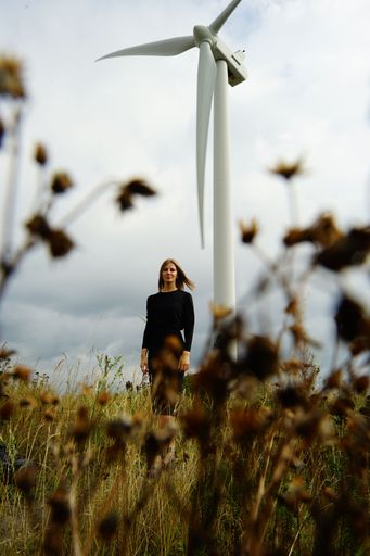A young woman posing in a field of wild grass and shrubs, with a large wind turbine in the distance