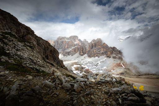 A scenic shot of a high rocky peak under the cloudy blue sky with rocks and stone on its foot