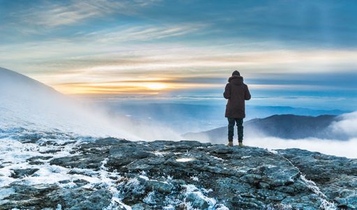 A person standing on a snow covered cliff over the breathtaking view of the mountains under the sunset