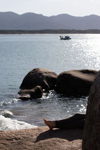 A vertical of a person's legs on a sandy beach with rocks and a boat in the background