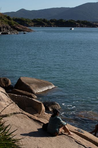 A vertical of a woman sitting on a rocky beach on a sunny day