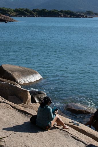 A vertical of a woman sitting and checking her phone on a rocky beach on a sunny day