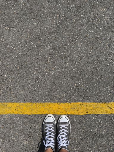 A vertical shot of a man's black sneakers next to an orange line on an asphalt road