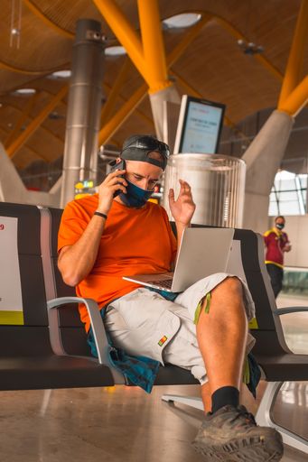 A vertical shot of a young man working on a laptop in an airport wearing a sanitary mask