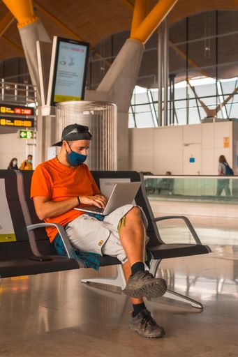 A vertical shot of a young man working on a laptop in an airport wearing a sanitary mask