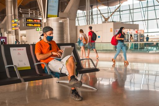 A closeup shot of a young man working on a laptop in an airport wearing a sanitary mask