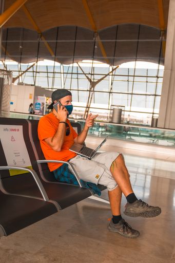 A closeup shot of a young man working on a laptop and talking in an airport wearing sanitary mask