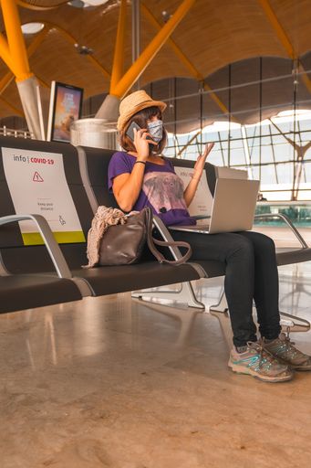 A vertical shot of a female in a protective face mask talking on her mobile phone and using her laptop while sitting in the waiting zone of an airport