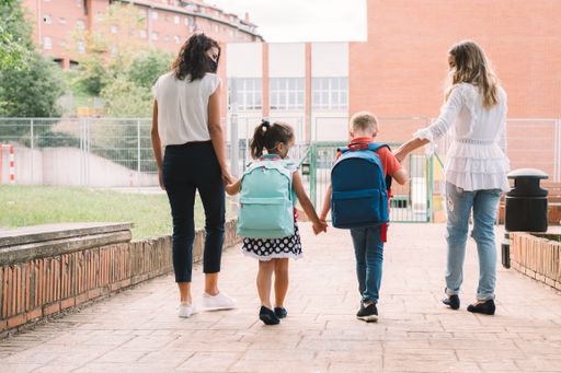 Two women and two children holding hands from back go to school for first time after coronavirus pandemic. Mothers accompanies the students along way.