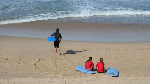 rear view of two teenage girls sitting next to blue surfboards on sand with teenage boy carrying surfboard walking towards water at the beach