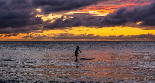 A dramatic view of a colorful sunset over the sea with the silhouette of a surfer