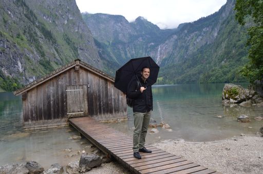 An elegant caucasian man with an umbrella standing on a wooden dock leading to a log cabin in Berchtesgaden National Park