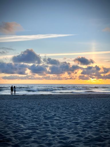 A vertical shot of a couple standing at the beach and watching the sunset behind the cloudy sky