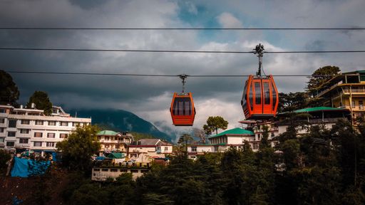 The orange cable cars next to the colorful buildings surrounding trees with rainy clouds in the background, Himachal Pradesh India