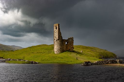 A scenic view of Ardvreck Castle on a green hill against a lake on a cloudy day in Sutherland, Scotland